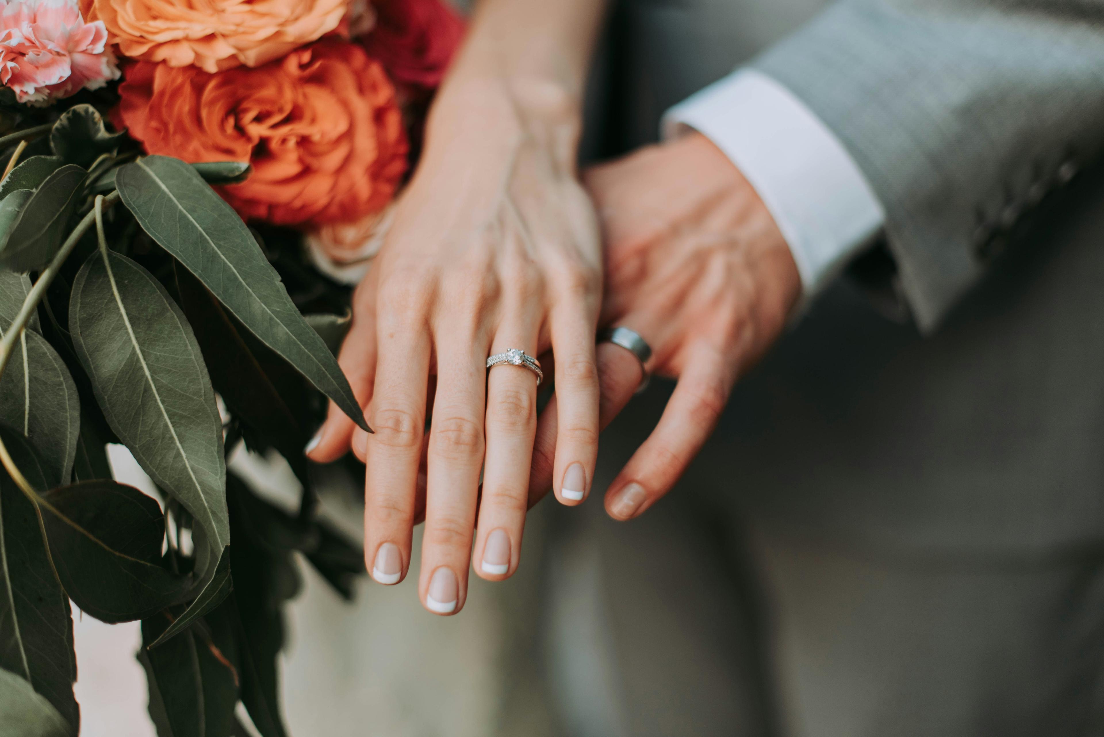 Couple's hands showing wedding rings beside a bouquet of orange roses