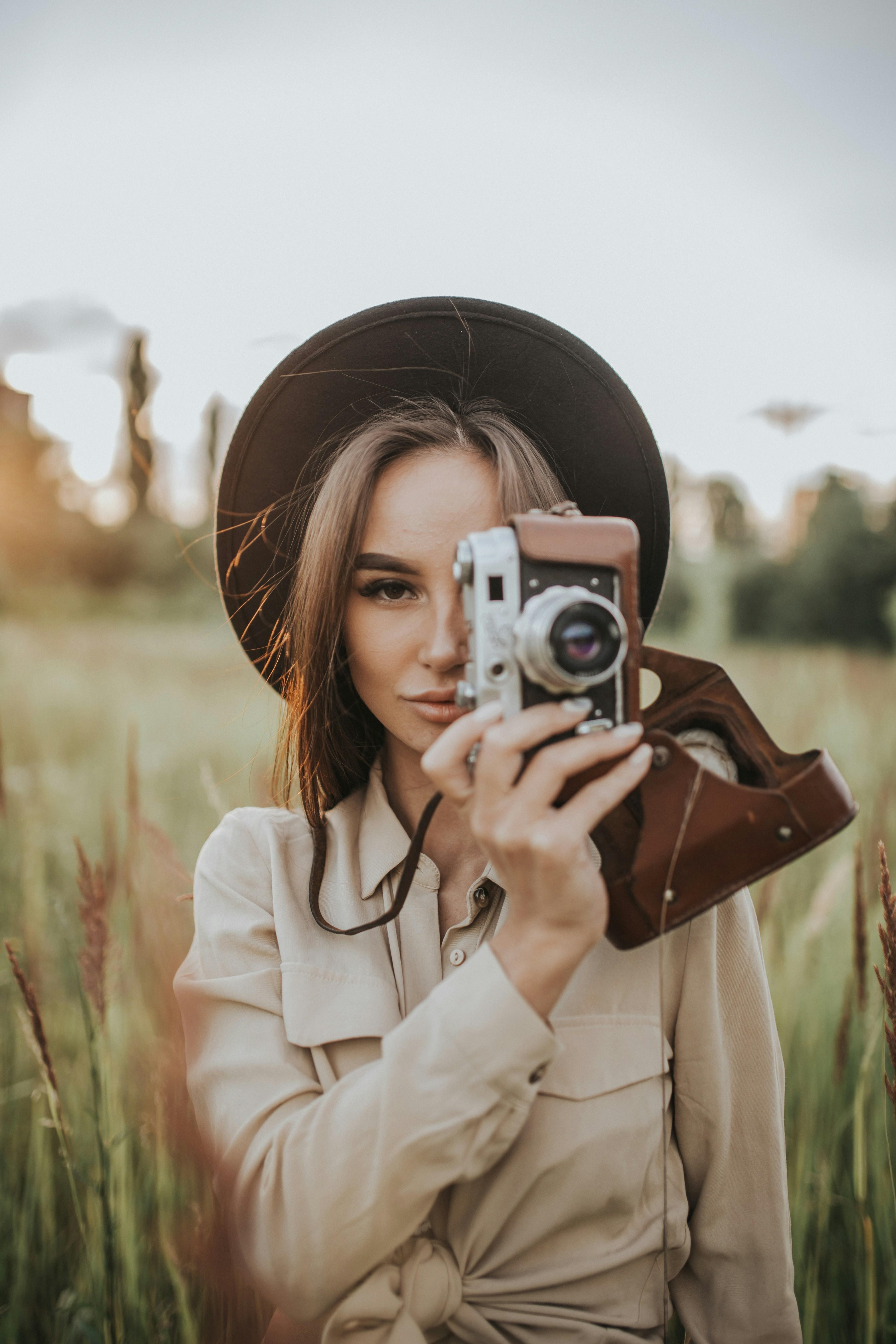 Woman in a wide-brim hat holding a vintage film camera in a grassy field