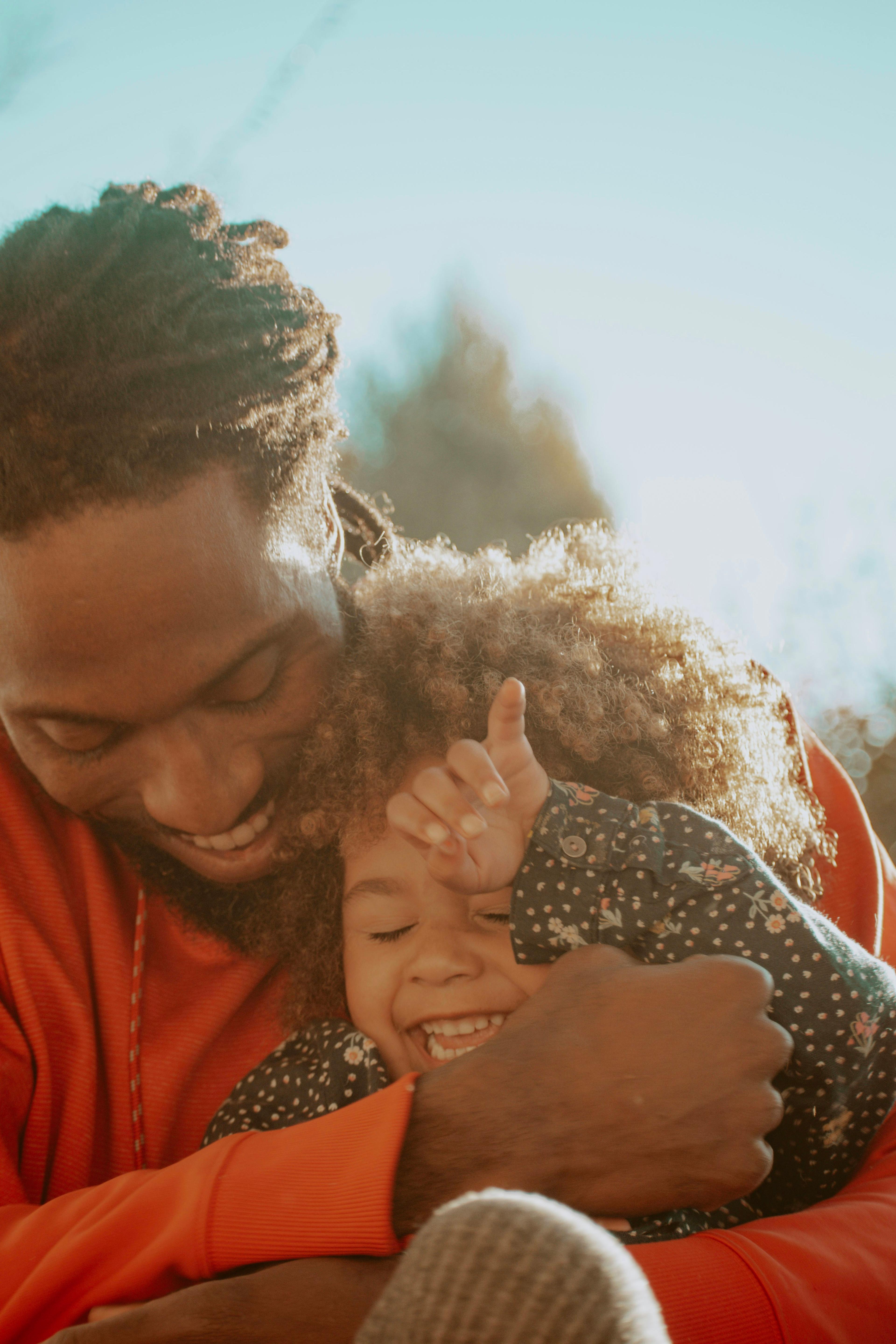 Father in an orange sweater hugging his laughing young daughter outdoors in warm sunlight