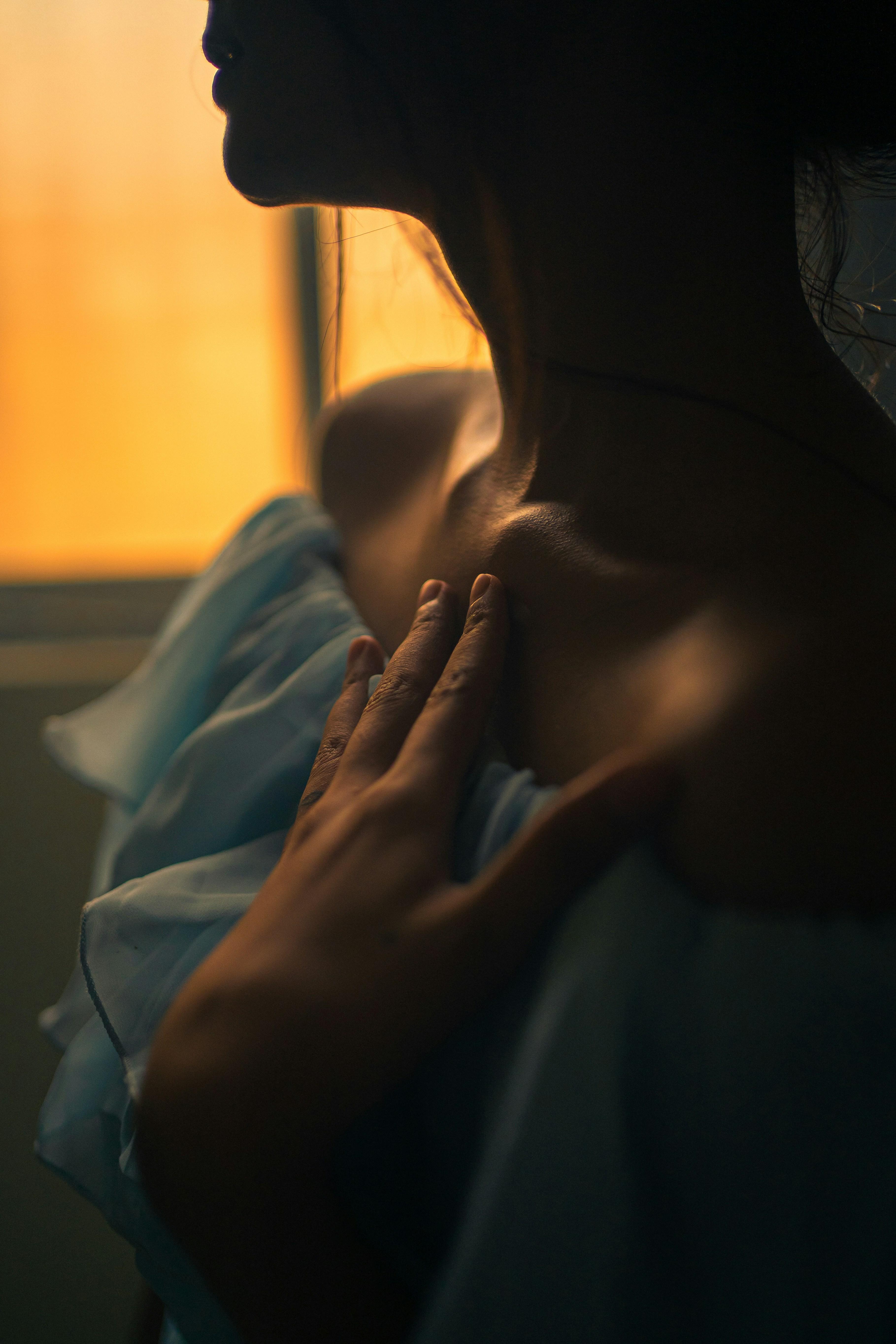 Close crop of a woman's neck and shoulder with her hand on her chest by a sunlit window